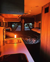Cozy RV rental interior at night, showcasing a kitchen sink, bed, & warm ambient lighting with street view.