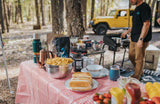 RV rental adventure: A man cooks a delicious outdoor meal at a vibrant campsite, with hot dogs and snacks on a checkered table.