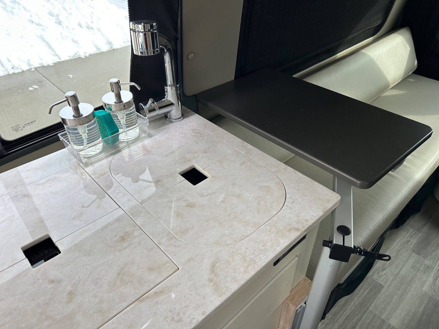 Inside an RV rental: a modern kitchen with a light-colored counter, sink, faucet, and a dark table by a cream bench.