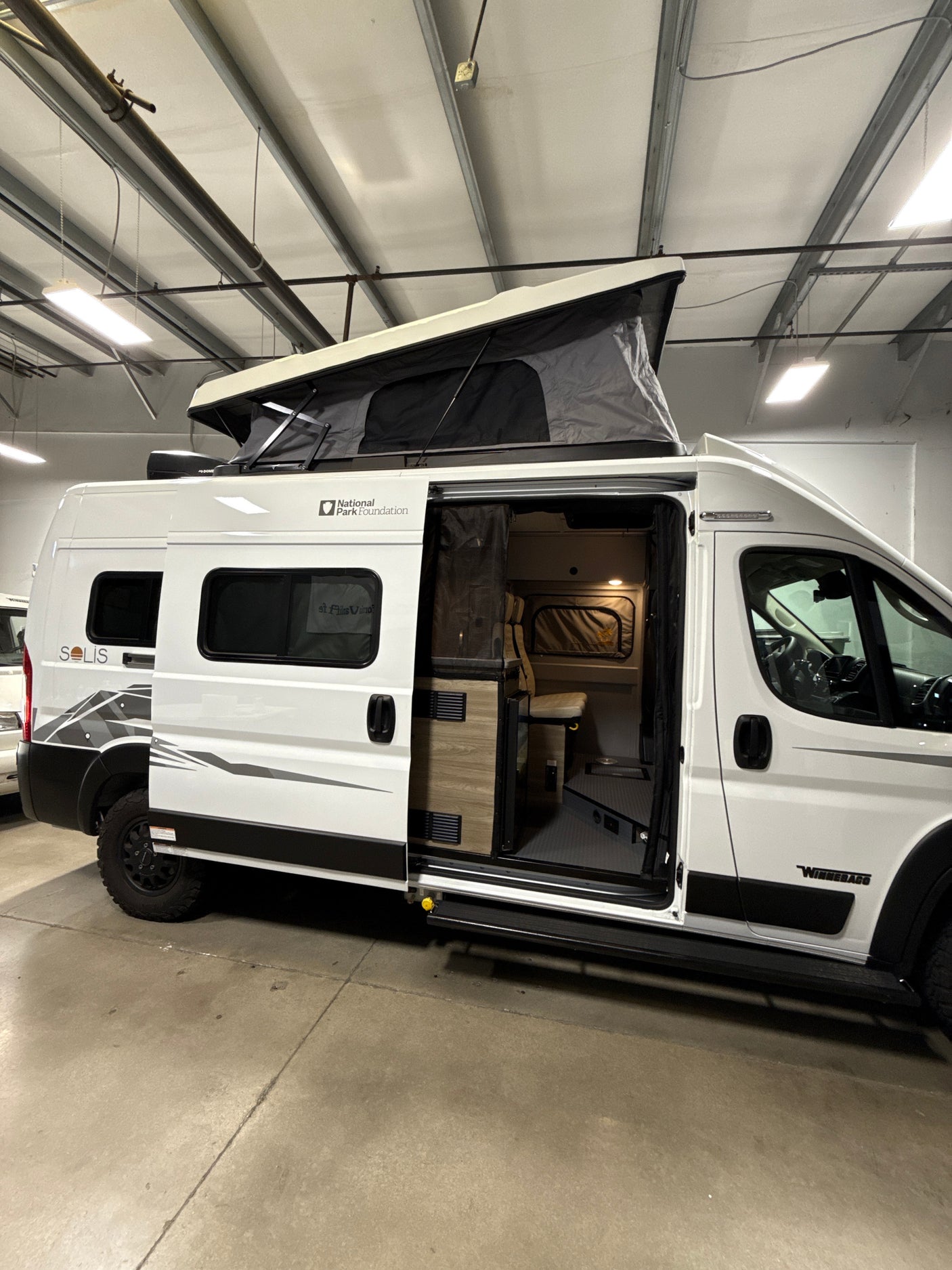 White Winnebago Solis camper van with open pop-top tent and interior, parked indoors. Ideal for your next RV rental trip.