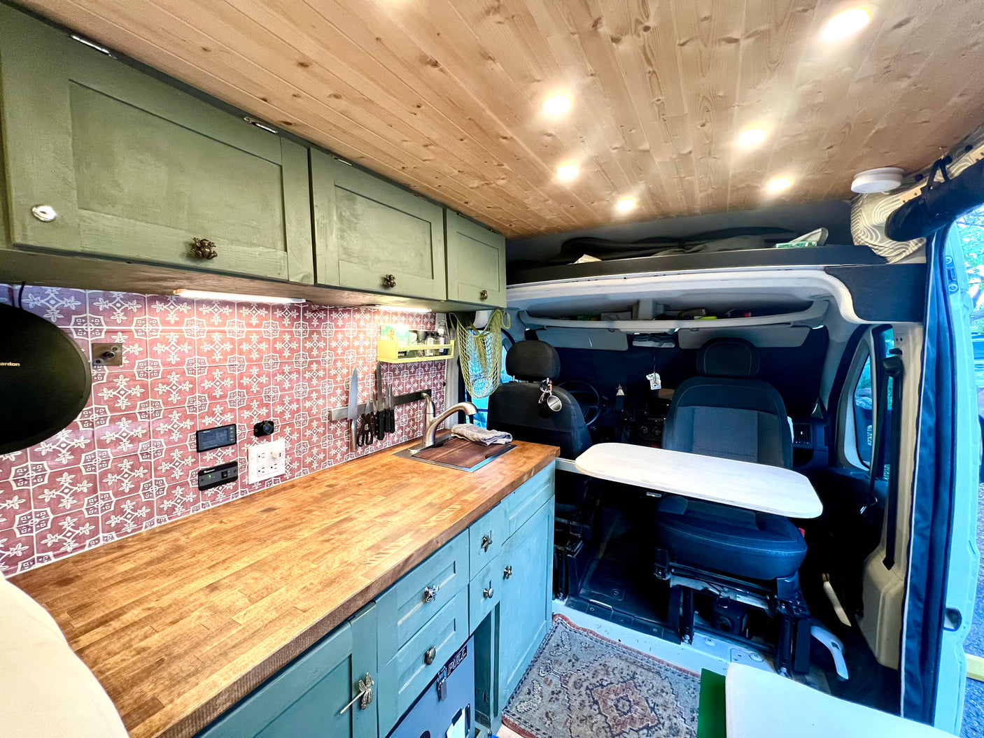 Cozy rv rental campervan kitchen with wooden ceiling, green cabinets, unique backsplash, and front seats with a swivel table.