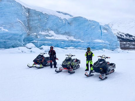 Three snowmobilers pose by a massive blue glacier. Your next winter adventure, accessible with an RV rental, awaits!