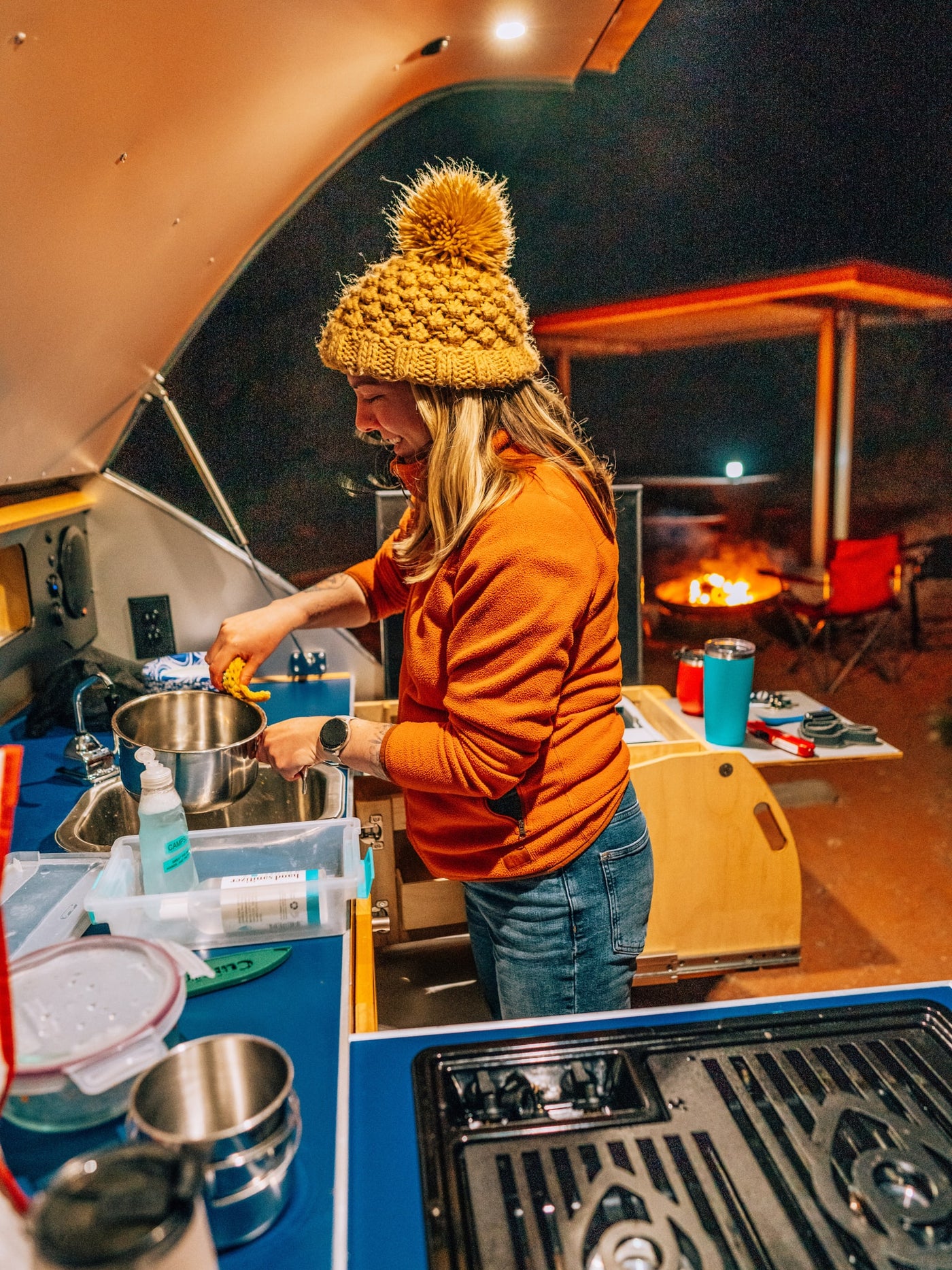 A woman washes dishes at the compact outdoor kitchen of an RV rental, a cozy campfire glowing in the background under the night sky. — photo 3