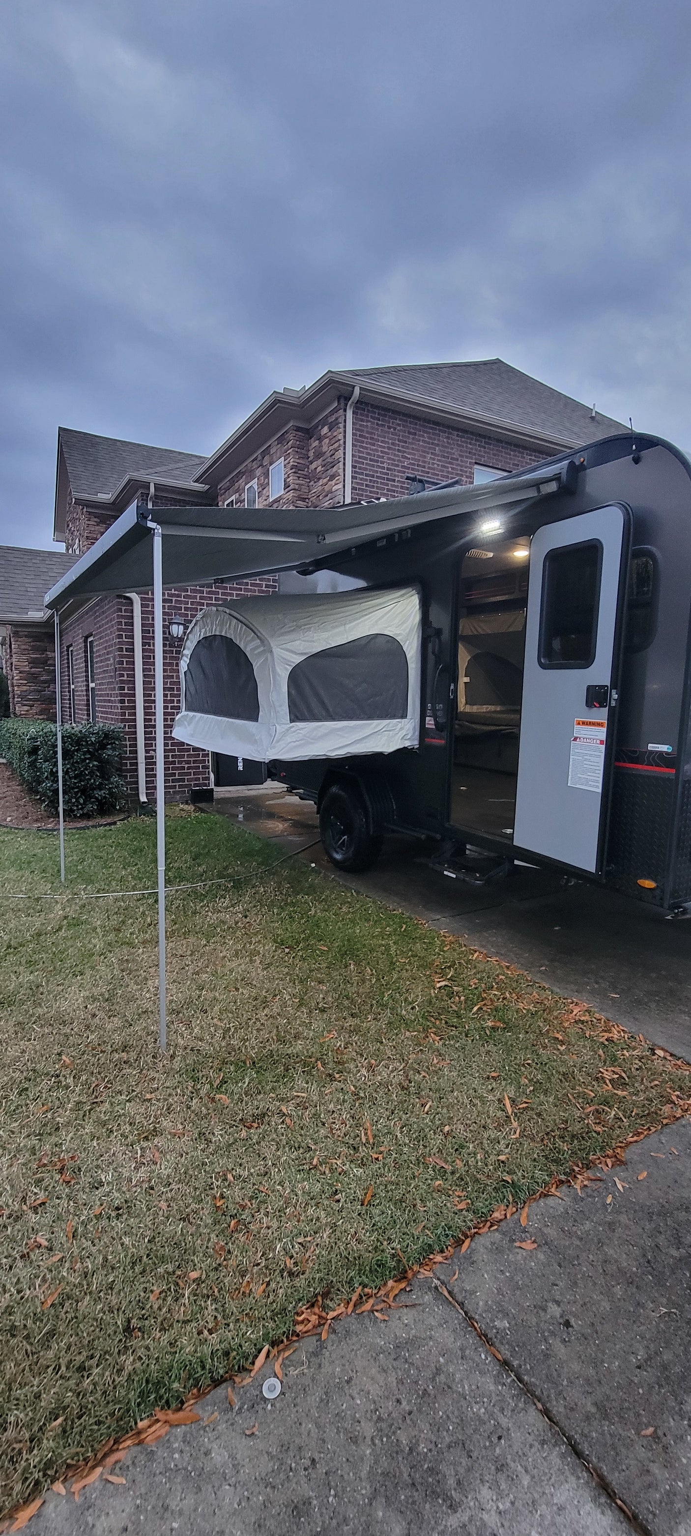 Modern popup camper with an extended awning and pop-out bed, ideal for an rv rental. Parked on a driveway next to a brick house.