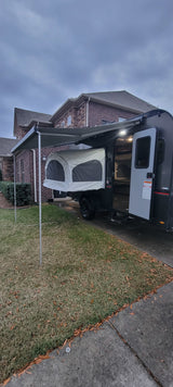 Modern popup camper with an extended awning and pop-out bed, ideal for an rv rental. Parked on a driveway next to a brick house.