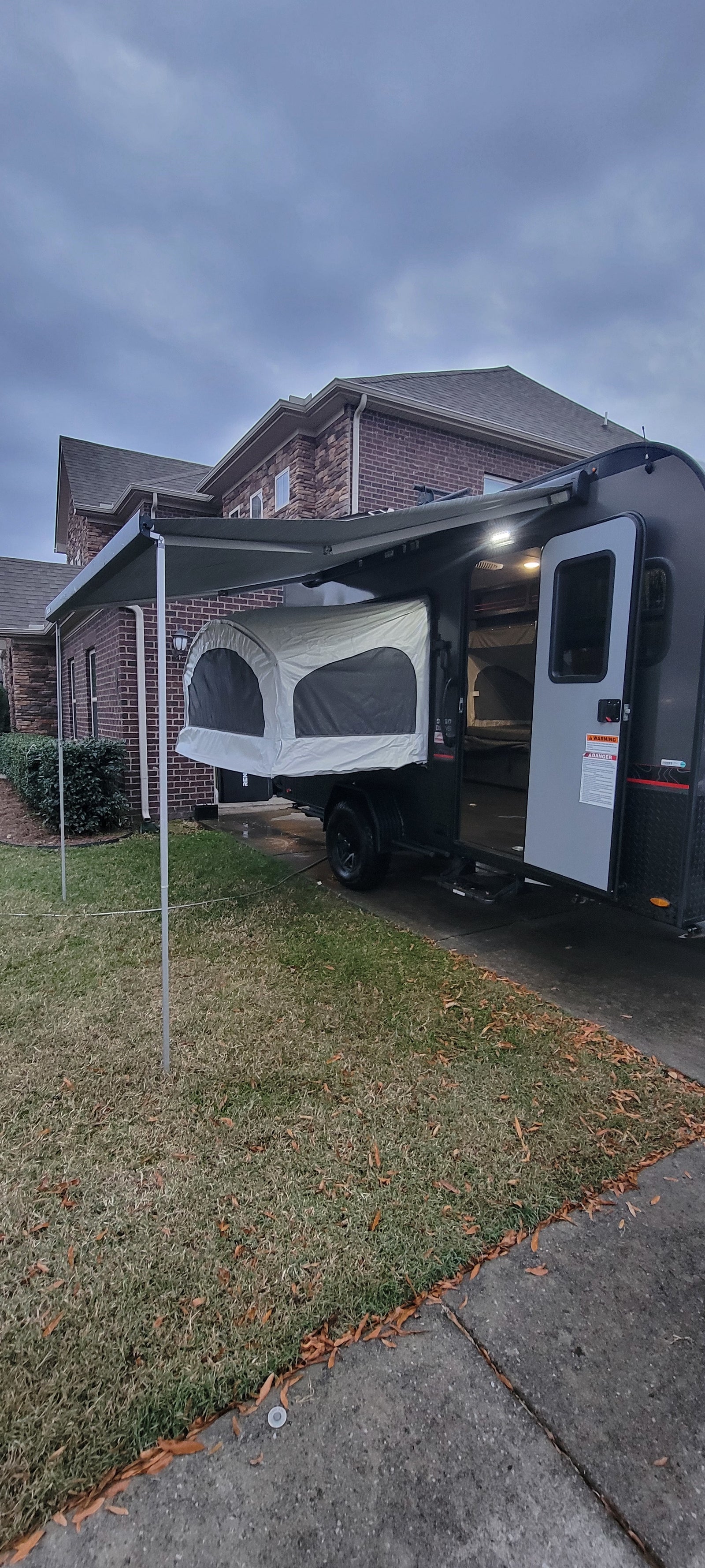 Modern popup camper with an extended awning and pop-out bed, ideal for an rv rental. Parked on a driveway next to a brick house.