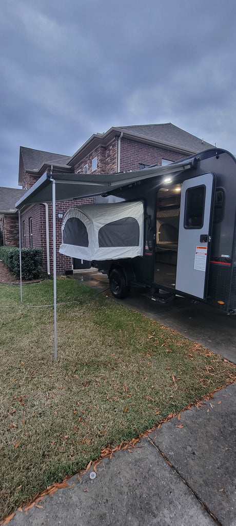 Modern popup camper with an extended awning and pop-out bed, ideal for an rv rental. Parked on a driveway next to a brick house.