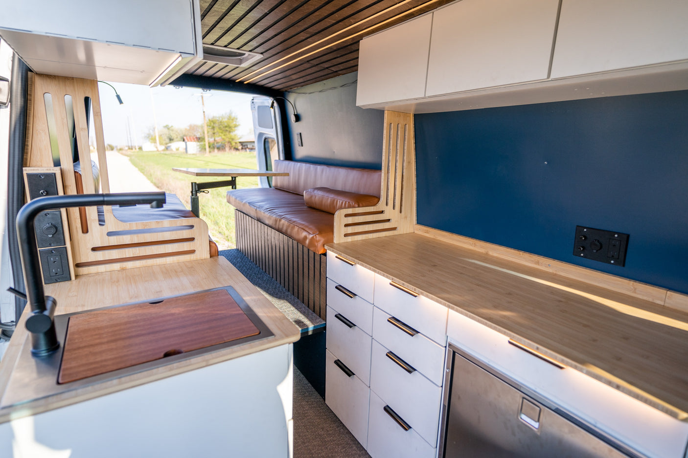 Inside a modern RV rental: a bright bamboo kitchen with sink, comfy brown bench seating, and smart storage units.