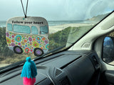 RV rental view: A 'Follow your heart' van ornament hangs as a serene beach and ocean stretch beyond the windshield. Van life.