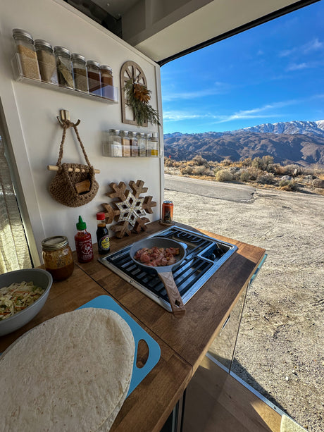 RV rental cooking setup: fresh meal on stove, sauces, spices, and tortillas, with stunning mountain backdrop.