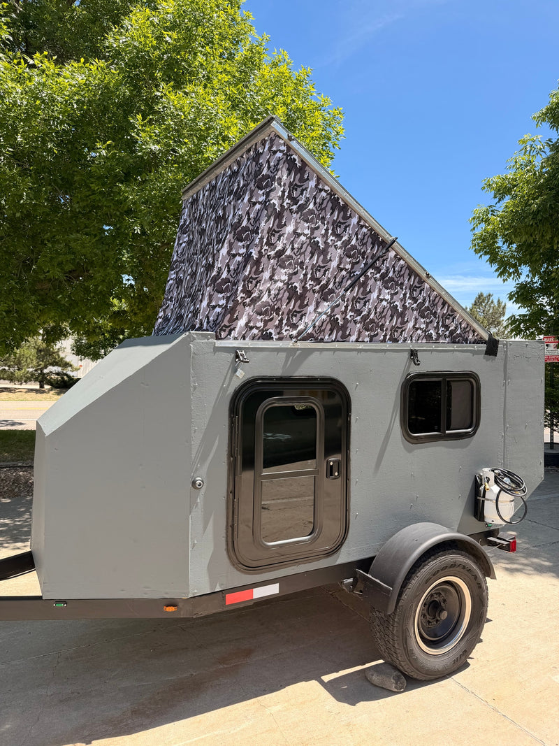 Compact grey camper trailer with camo pop-up roof, ideal for rv rental adventures. Features a door, window, and propane tank. — photo 1