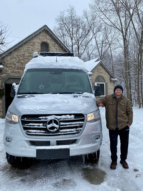 Man smiles next to his snow-covered Mercedes-Benz RV rental van, ready for winter travel adventures.