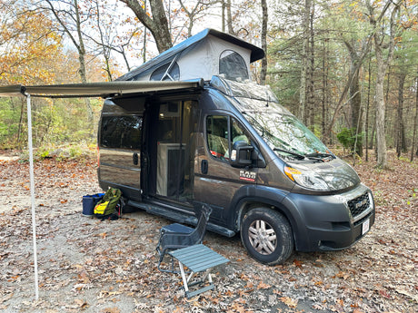 Grey Ram ProMaster RV rental with pop-top extended & awning out, parked in a scenic autumn forest campsite.