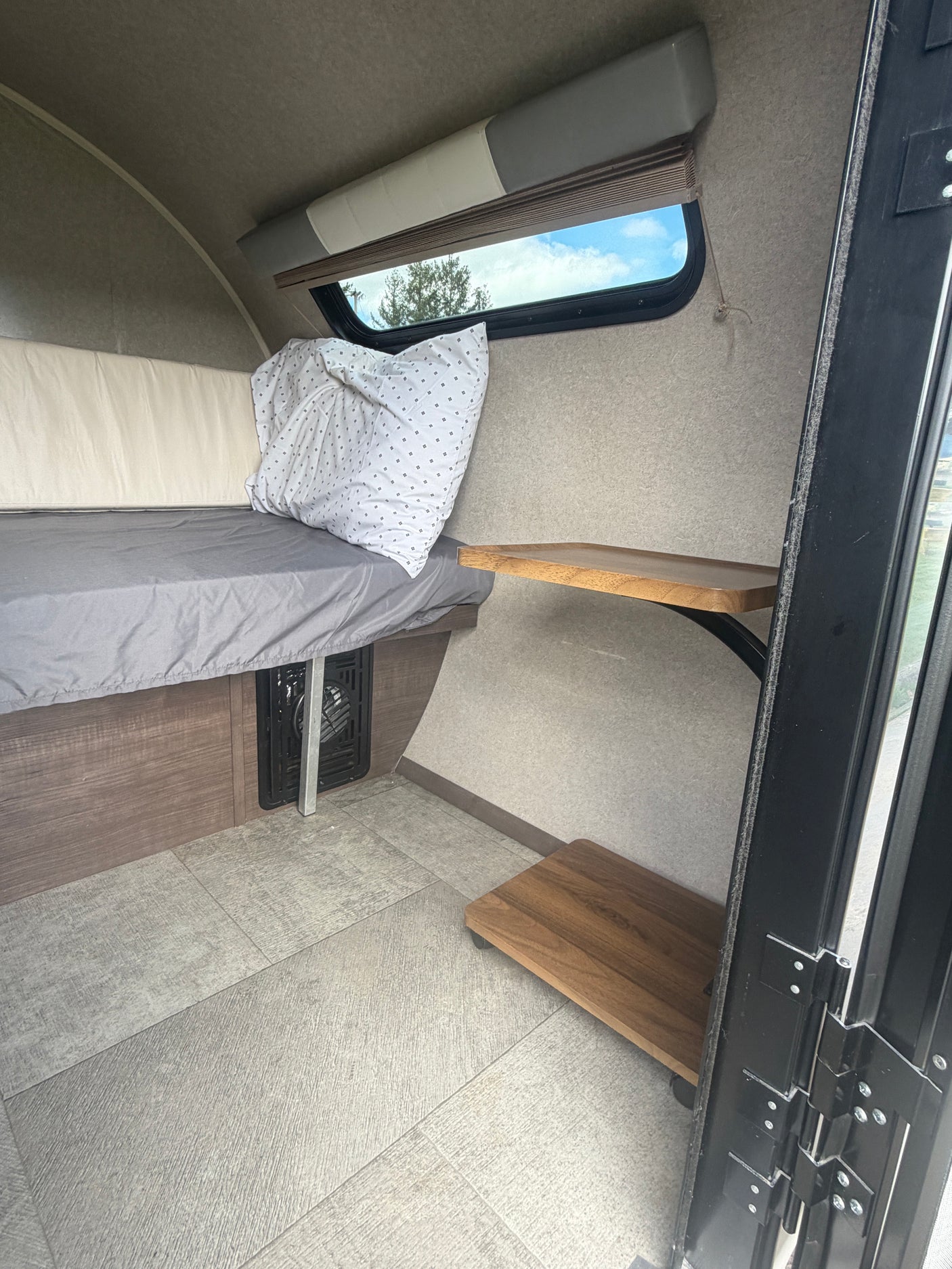 Cozy RV rental interior with a bed, grey sheet, window view of trees, and handy wooden shelves for minimalist travel.