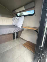Cozy RV rental interior with a bed, grey sheet, window view of trees, and handy wooden shelves for minimalist travel.