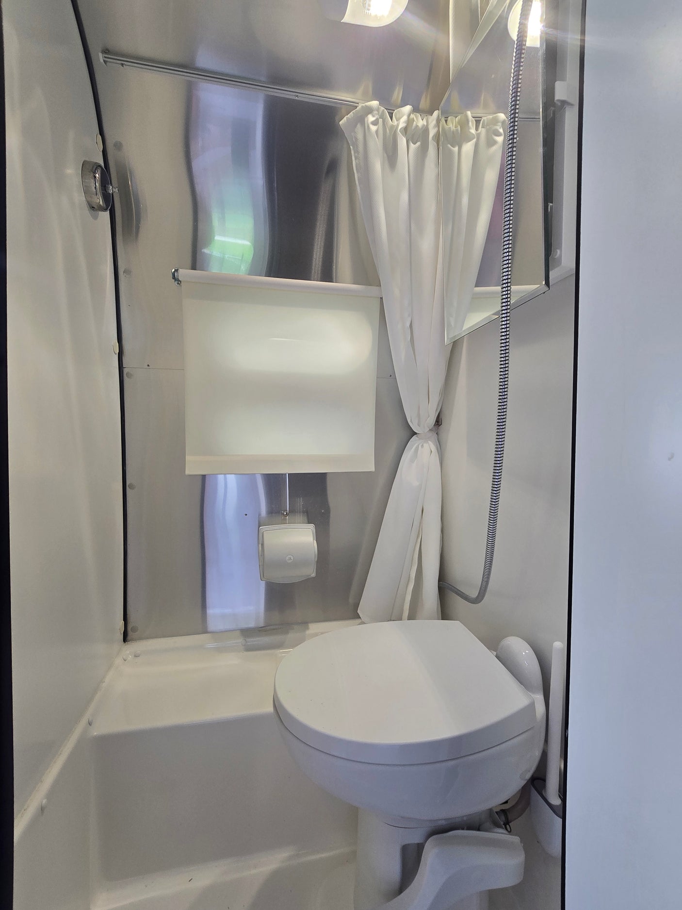 Compact RV rental bathroom featuring a toilet, shower with curtain, and mirrored cabinet on metallic walls, optimizing space.