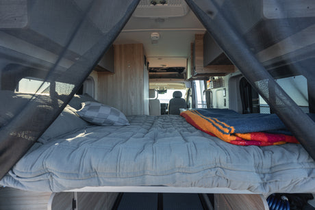 RV rental: Cozy camper van interior featuring a comfy grey bed with a colorful blanket, looking through netting to the front cab.