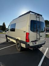 Light-colored Mercedes Sprinter camper van with roof rack, ladder, and AWD, parked outdoors. Perfect for rv rental adventures.