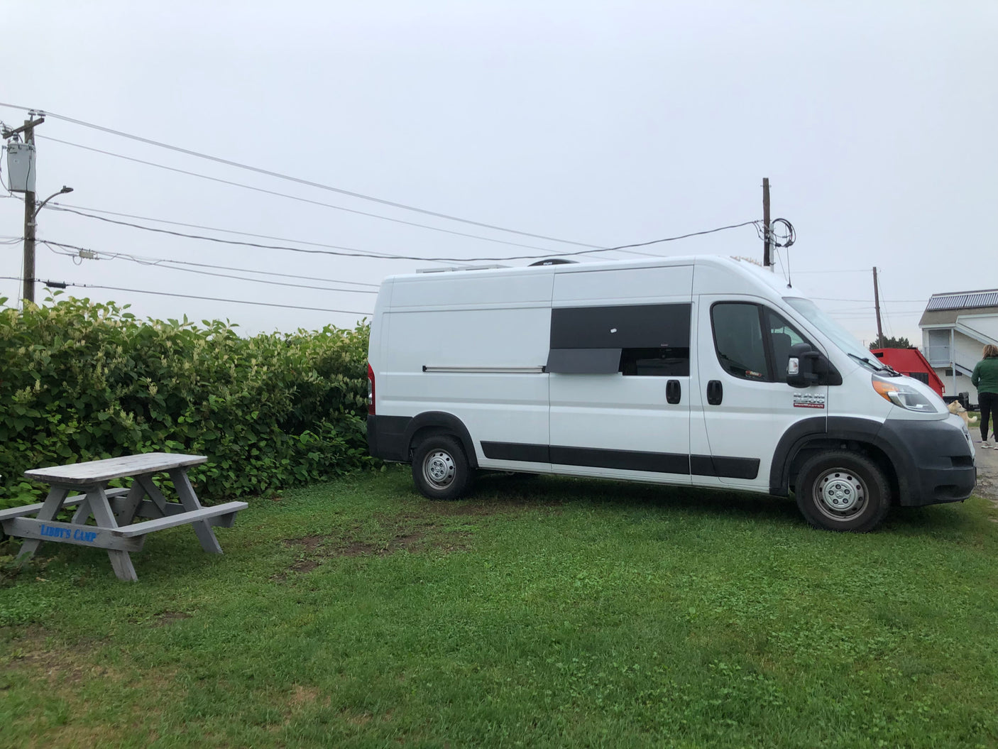 A white campervan is parked on grass next to a picnic table. This RV rental is ready for adventure under an overcast sky.