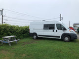 A white campervan is parked on grass next to a picnic table. This RV rental is ready for adventure under an overcast sky.