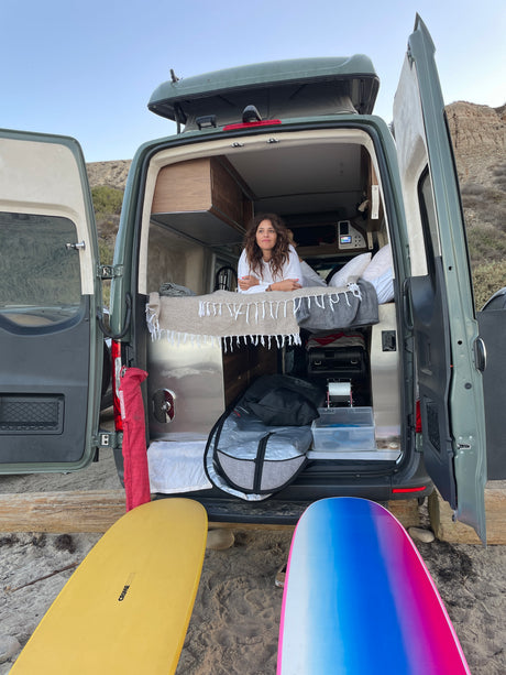 Coastal RV rental: Woman relaxing in a camper van at the beach, surfboards in foreground, cliff background.