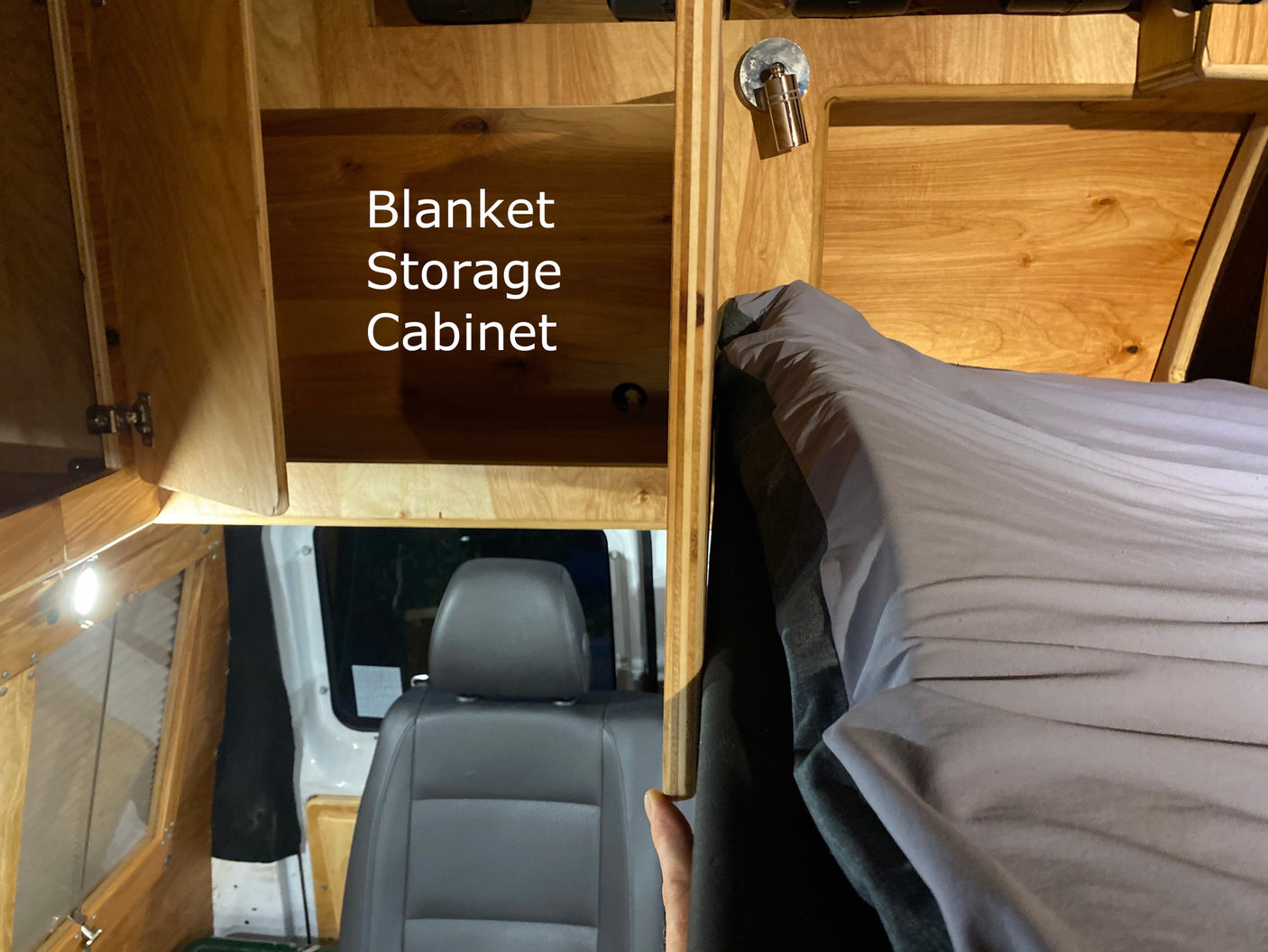 Inside an RV rental: a wooden blanket storage cabinet, cozy bed with gray linen, and vehicle seat visible in this camper van interior.
