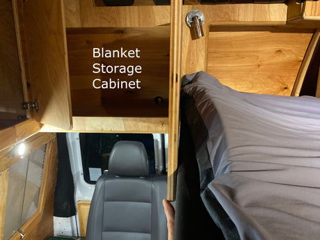 Inside an RV rental: a wooden blanket storage cabinet, cozy bed with gray linen, and vehicle seat visible in this camper van interior.