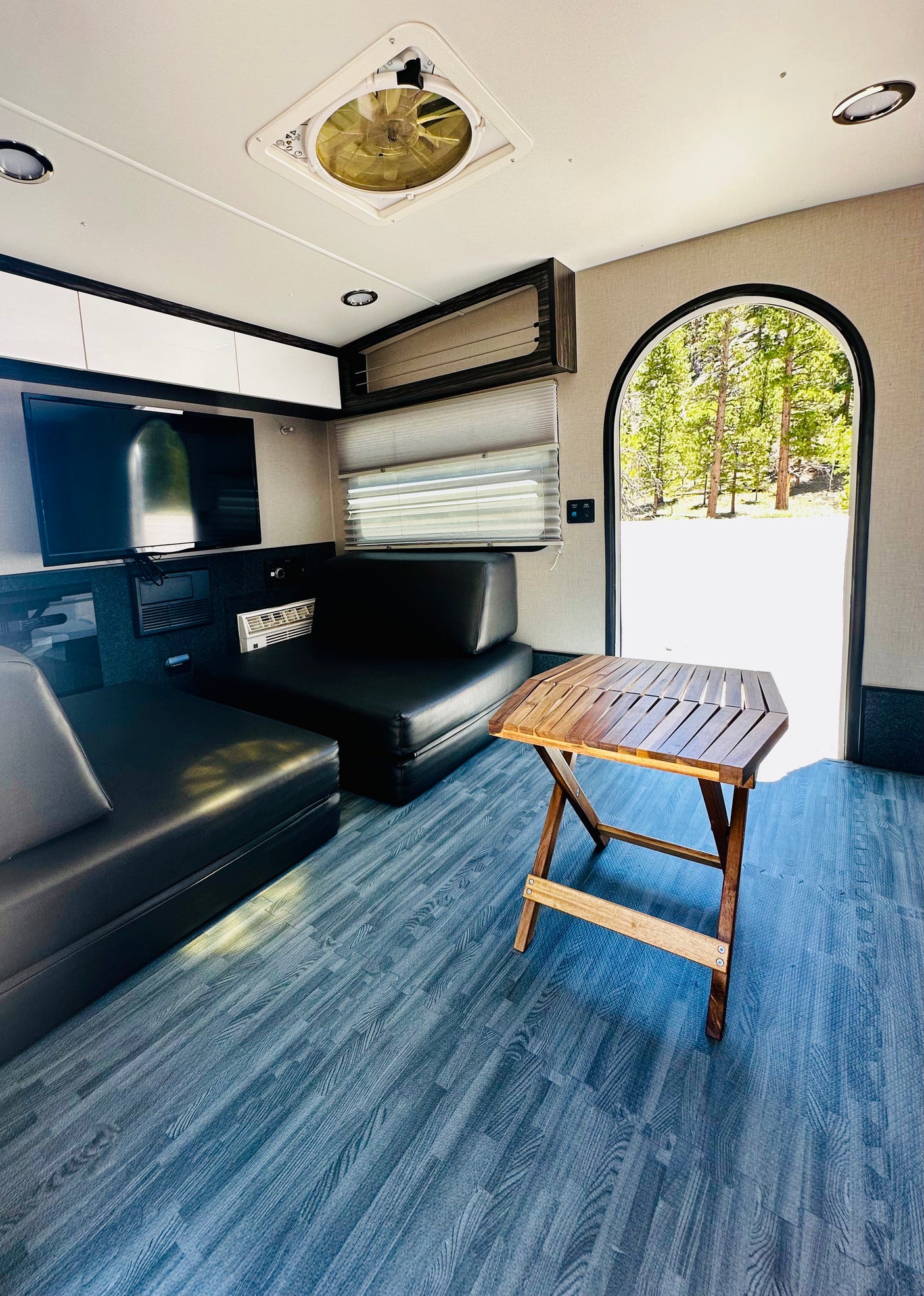 Modern RV rental interior: black seating, folding wood table, blue floor. Arched door views to nature & forest.