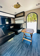 Modern RV rental interior: black seating, folding wood table, blue floor. Arched door views to nature & forest.