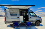 Custom gray camper van rv rental set up for adventure on a salt flat, with open door, extended awning, chairs & table.