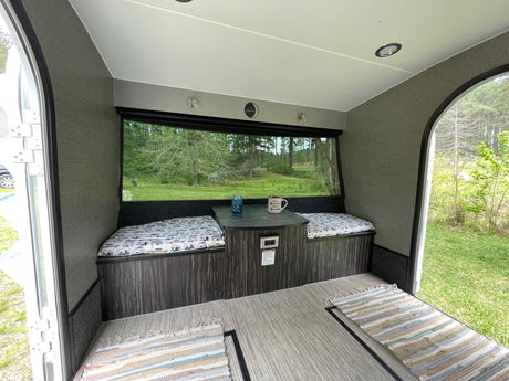 Interior of an RV rental featuring a cozy dinette with cushioned benches, a table, and a large window showcasing a forest view.