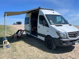 Relaxing under the awning of a white Mercedes Sprinter RV rental van, set up for camping with cooking gear.