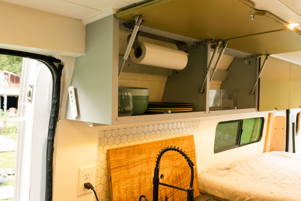 RV rental kitchen showing open overhead cabinets with dishes, paper towels, a sink with a gooseneck faucet, and hex tile backsplash. — photo 11