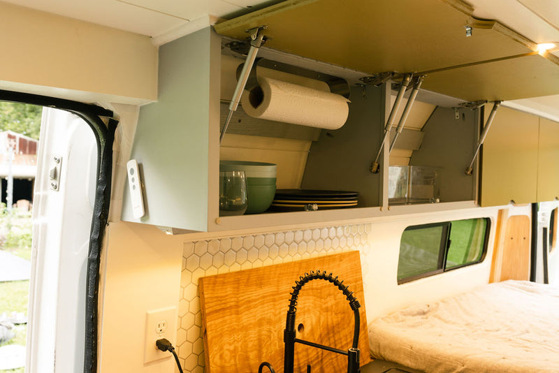 RV rental kitchen showing open overhead cabinets with dishes, paper towels, a sink with a gooseneck faucet, and hex tile backsplash. — photo 11