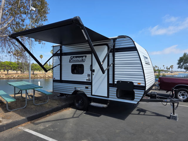 A white Coleman travel trailer RV rental with an extended black awning sits next to a lake