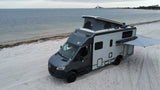 A grey and white RV rental is parked on a wide sandy beach by the ocean under a cloudy sky, with a long bridge in the distance.