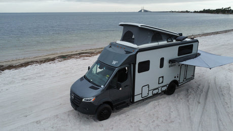 A grey and white RV rental is parked on a wide sandy beach by the ocean under a cloudy sky, with a long bridge in the distance.