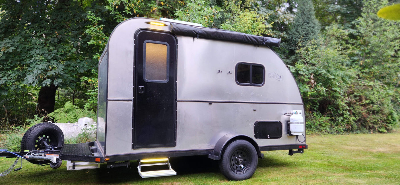 Silver teardrop RV rental parked on grass amidst trees, featuring a door, windows, spare tire, and a propane tank.