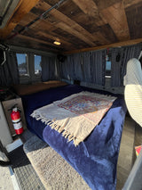 Inside a cozy RV rental: a plush bed, wooden ceiling, fishing rods, and patterned curtains create a warm van conversion.