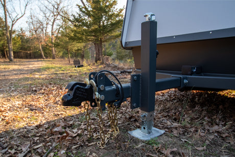 An RV rental unit's front hitch and deployed jack stand are visible, parked on a natural, leafy forest floor.