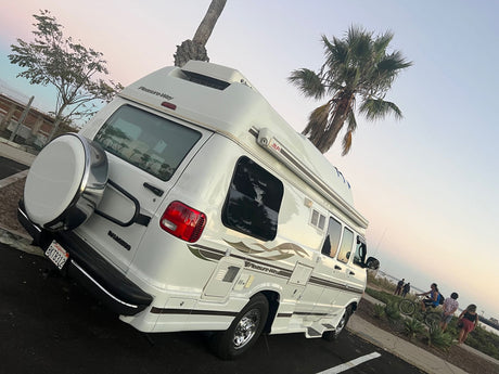 White Pleasure-Way RV rental parked by the water with palm trees & people. Perfect for a scenic adventure!