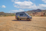 Adventure-ready gray rv rental van with yellow accents in a dirt lot, backed by scenic mountains and blue sky.