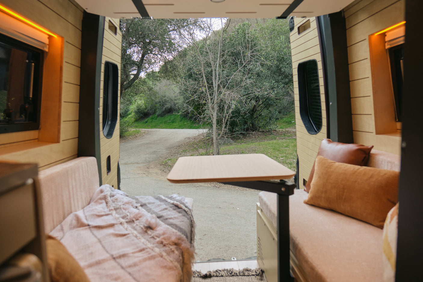 Cozy RV rental interior with wood paneling, seating, and a table, offering a serene view of a dirt road and nature.