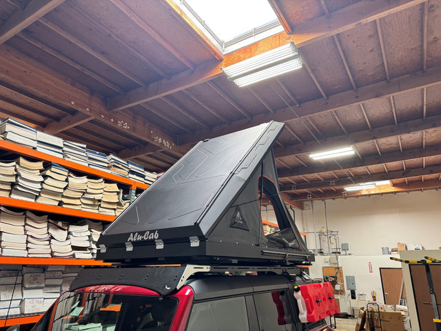 Alu-Cab Gen 3-R hardshell rooftop tent mounted on a roof rack above a red SUV, shown fully opened in its wedge configuration with the aluminum shell raised, Alu-Cab logo on the side panel and the mesh door panel visible
