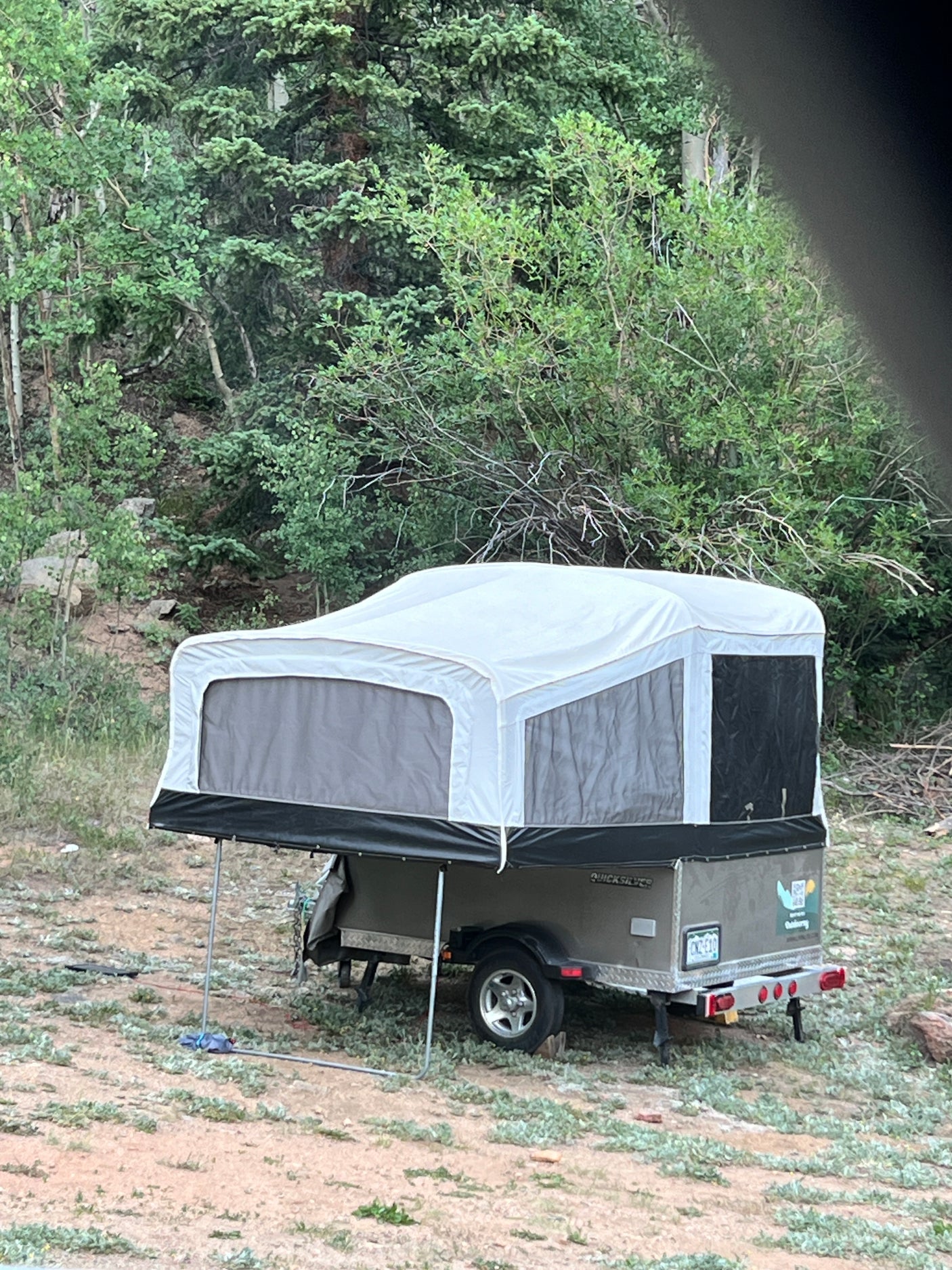 A grey Quicksilver pop-up camper tent trailer is set up in a wooded campsite, ready for your RV rental outdoor adventure.