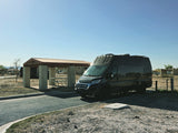 Modern dark grey campervan, popular for rv rental, parked at a dry campsite with covered picnic area. Sunny sky.