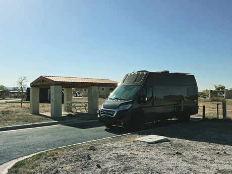Modern dark grey campervan, popular for rv rental, parked at a dry campsite with covered picnic area. Sunny sky.
