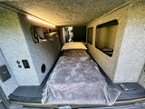 Inside an RV rental: a single bed with pillow & blanket, overhead lighting, and carpeted walls with built-in storage.