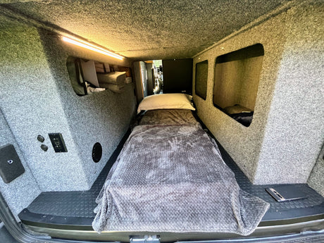 Inside an RV rental: a single bed with pillow & blanket, overhead lighting, and carpeted walls with built-in storage.