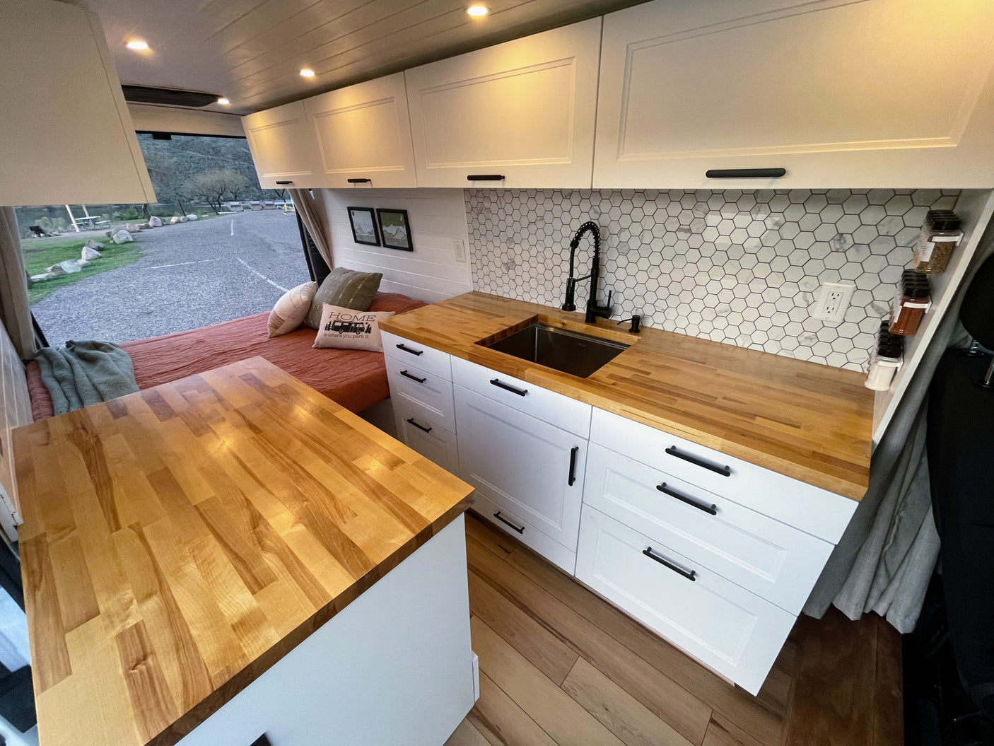 Spacious RV rental interior featuring a modern kitchen with wood counters, white cabinets, hex backsplash, and a cozy bed area.